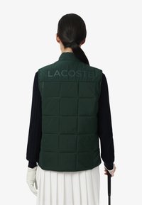 Groene puffer gilet met gestikt patroon en hoge kraag. Heeft grote "LACOSTE" branding op de achterkant. Glad materiaal, mouwloos ontwerp.