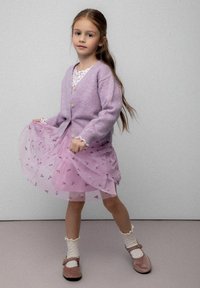 Fioletowy kardigan z okrągłymi guzikami, warstwowa różowa spódnica tiulowa z kwiatową haftem, białe skarpetki z falbankami i brązowe buty.