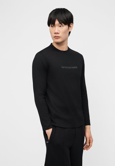 Νέος άνδρας που φοράει μαύρο μακρυμάνικο πουκάμισο με το λογότυπο "Emporio Armani", στέκεται με το ένα χέρι στην τσέπη, κοιτάζοντας προς τα αριστερά του.