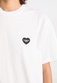 T-shirt en coton blanc avec un patch en forme de cœur noir portant le texte "Carhartt WIP", encolure ronde et manches courtes.