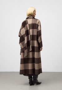 Cappotto lungo e sovradimensionato in motivo a scacchi marrone e beige, realizzato in un morbido materiale simile alla lana. Tessuto aggiuntivo drappeggiato su una spalla.