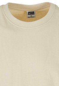 Tricou din bumbac de culoare crem, cu guler rotund, mâneci scurte și un singur etichetă care arată "URBAN CLASSICS M, Fabricat în Bangladesh."