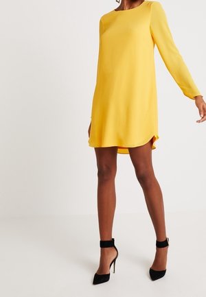 Mujer vestida con un mini vestido amarillo de manga larga con bajo redondeado y zapatos de tacón alto con correa al tobillo negra, de pie frente a un fondo liso.