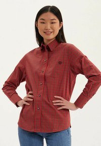 Chemise à carreaux rouge et noire à manches longues, avec col, boutons contrastants et un petit logo sur la poitrine.