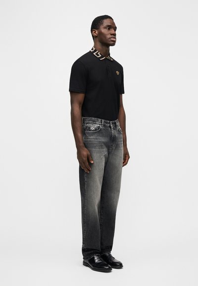 Juoda polo marškinėliai su raštuotu apykakliu, pilki išblukę džinsai ir juodi batai. Marškinėliuose yra logotipas, o džinsuose – detalės.