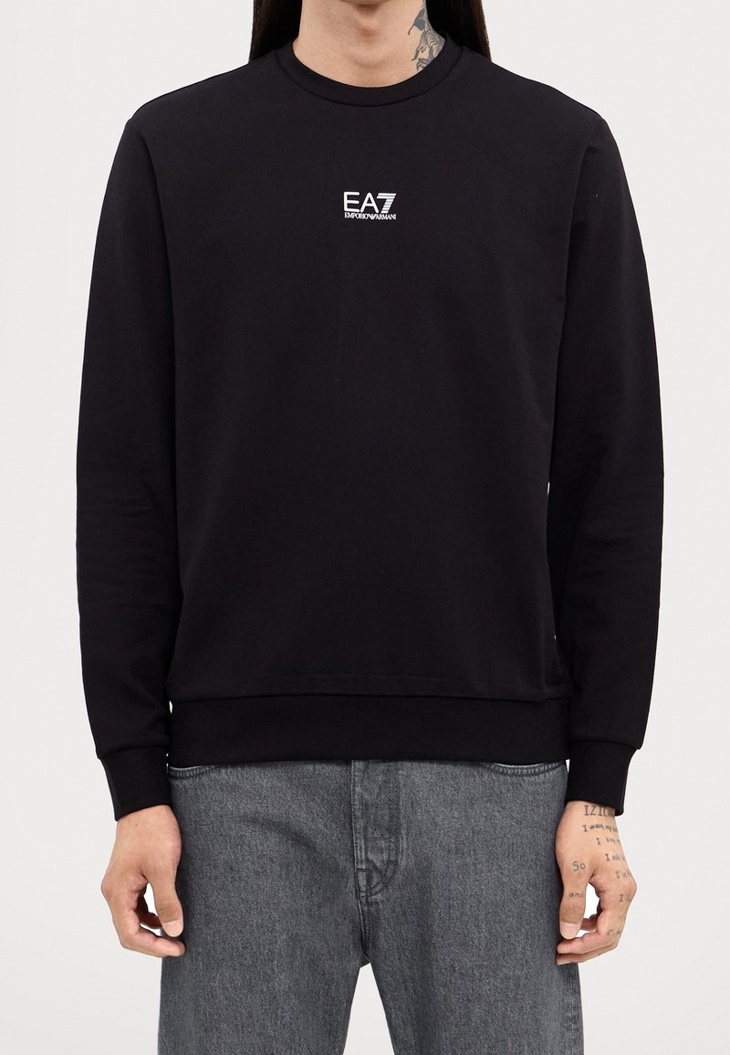 Personne portant un sweat-shirt noir avec le logo "EA7 Emporio Armani" sur la poitrine et un jean gris, tatouages visibles au cou et aux mains.