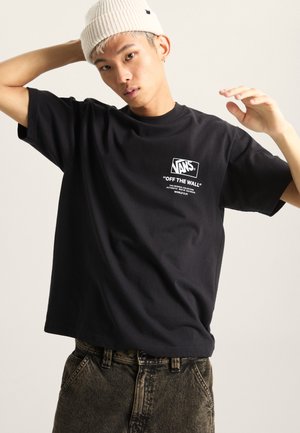 Svart bomulls t-shirt med korta ärmar, med en vit Vans-logotyp och slogan tryckt på bröstet. Bärs med mörka, slitna jeans och en beige beanie.