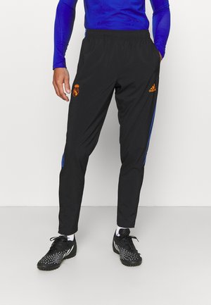 Personne portant un pantalon de survêtement noir Real Madrid Adidas avec des logos orange, une chemise à manches longues bleue, des chaussures de sport noires et des chaussettes blanches.