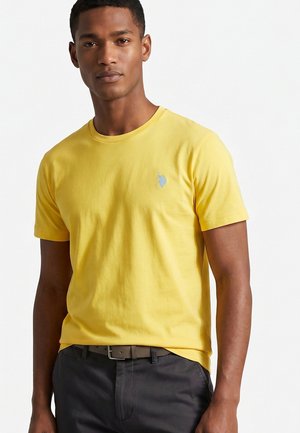 Jonge man die een effen geel T-shirt met korte mouwen draagt met een klein geborduurd logo op de borst en donkergrijze broek.