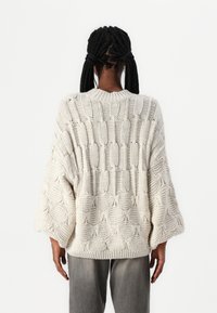 Personne aux cheveux tressés porte un pull tricoté crème surdimensionné avec des motifs torsadés et boules texturés, associé à un pantalon gris, vue de dos.