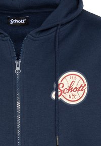 Sudadera con cremallera en color azul marino, hecha de una tela suave, que presenta un logo redondo en rojo y blanco con el texto "Schott NYC" y "1913". Incluye cordones en la capucha.