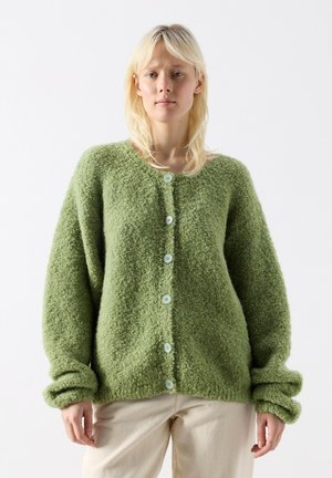 Žena s blond vlasmi, oblečená v voľnom zelenom zapínacom kardigane s textúrou a svetlo béžových nohaviciach, na jednoduchom bielom pozadí.