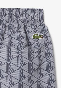 Tissu bleu clair à motif géométrique, taille élastique, détail de poche et logo crocodile vert brodé.