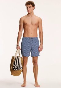 Męskie kąpielówki w kolorze niebieskim z białym wzorem fal, z elastycznym paskiem i sznurkiem do regulacji. Trzymana jest pleciona torba ze słomy z czarnymi paskami.