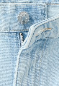 Jean en denim bleu clair partiellement dézippé, montrant une fermeture éclair métallique et un bouton argenté marqué du nom de la marque et de l'année.