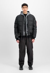 Joven de pie, vistiendo una chaqueta acolchada negra sobre una sudadera gris, pantalones cargo negros y zapatillas negras, contra un fondo claro y sencillo.