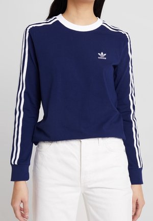 Personne portant un maillot à manches longues Adidas bleu marine avec des bandes blanches sur les manches et un pantalon blanc sur un fond uni.