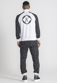 Chándal deportivo con una parte superior blanca que presenta un logo negro, y pantalones negros con un patrón repetitivo. Puños y tobillos elásticos.