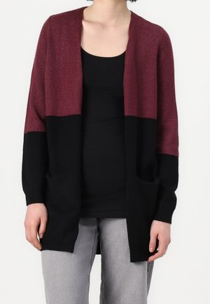 Persoon draagt een tweekleurig openvallend vest, met een bordeauxrode bovenkant en zwarte onderkant, over een zwart shirt en lichtgrijze broek.