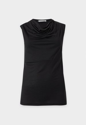 Vrouw staand met één hand in de zak, draag een mouwloze zwarte top, lichtblauwe jeans met wijde pijpen en zwarte puntige hakken op witte achtergrond.