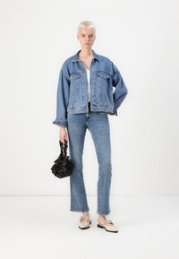 Giacca di jeans azzurro chiaro con una vestibilità rilassata, abbinata a jeans coordinati. Tiene in mano una borsa nera a fantasia; indossa scarpe beige slip-on.