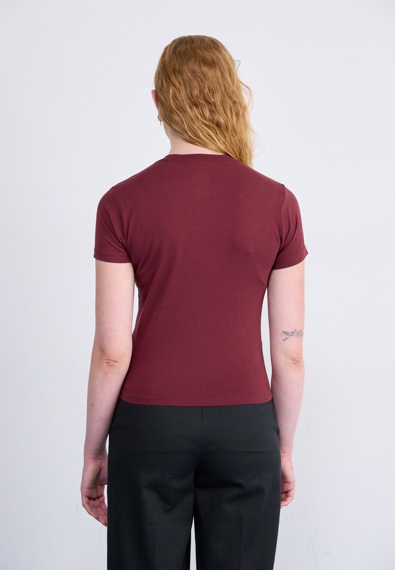 T-shirt ajustée bordeaux à manches courtes et col rond, associée à un pantalon noir. Texture lisse, design minimal, sans motifs visibles.