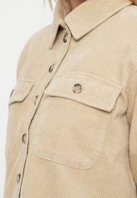 Μπεζ πουκάμισο από κορντούρα με μυτερό γιακά, δύο τσέπες στο στήθος και μεγάλους κουμπιά, που εμφανίζει υφή με ρίγες σε όλη του την επιφάνεια.