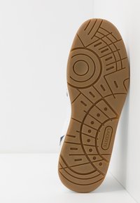 Vit sneakersula med en texturerad, gummiliknande yttersula som har cirkulära och linjära mönster, präglad logotyp och mörkblå detaljer.