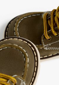 Chaussures en cuir marron avec des lacets jaunes, présentant des accents beige, des coutures blanches et des bouts ronds. Vue en gros plan mettant en avant les détails du matériau et du design.