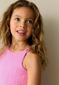 Roza obleka z teksturo, ki ima tanke naramnice in okrogel izrez. Otrok ima dolge kodraste lase in se smeji z vidnimi zobmi.