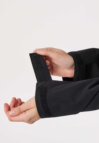 Χέρια που ρυθμίζουν τη μανσέτα ενός μαύρου σακακιού με αυτοκόλλητο Velcro, πάνω σε απλό φωτεινό φόντο.