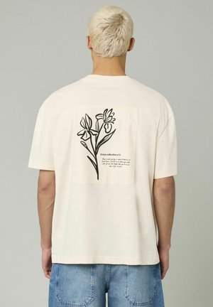 T-shirt à manches courtes de couleur crème présentant un imprimé floral noir au dos, avec un patch rectangulaire et du texte en dessous du design.