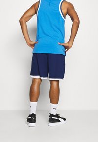 Man som står med händerna på höfterna, iförd en blå linne, marinblå shorts, vita Puma-strumpor och svart-vita Puma-sneakers, vänd bort.