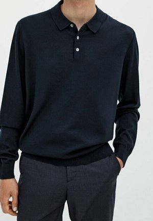 Uomo che indossa una polo blu navy a maniche lunghe con bottoni, infilata in pantaloni grigio scuro con una mano in tasca.