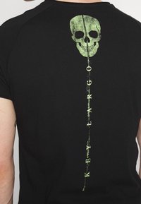 Svart t-shirt med en grön skallegrafik på baksidan, med vertikal text som läser "KEEP LARGO" under den. Korta ärmar och lätt tyg.