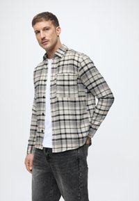 Koszula w kratę zapinana na guziki w kolorze beżowym i czarnym, z kieszonką na piersi, długimi rękawami i luźnym krojem, noszona na białym T-shircie.