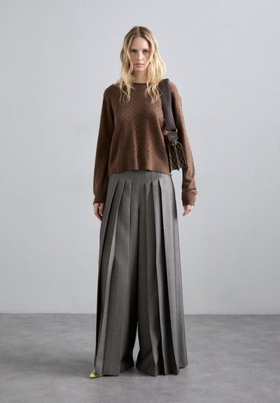 Kobieta w brązowym swetrze z ćwiekami, w szerokich szarych spodni plecionych, nosząca wzorzystą torbę na ramię, stojąca na szarej podłodze przed szarym murem.