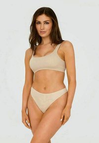 Kvinne med brunt hår som har på seg en beige ribbet en-skulder crop topp og matchende høyt skårne bikini-bukser, står mot en lys bakgrunn.