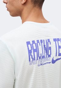 Νέος άνδρας φορεί λευκό αθλητικό πουκάμισο με τρύπες, με μεγάλο μπλε κείμενο "RACING TECH" και λογότυπο Nike στην πλάτη.