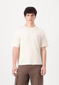 Camiseta de manga corta beige hecha de una tela suave, con un diseño bordado en azul claro en el lado izquierdo del pecho. Cuello redondo y corte relajado.