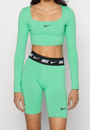Person trägt grünes langärmeliges Cropped-Top und passende grüne hoch taillierte Bike-Shorts mit schwarzen Nike-Logos auf Top und Bund.