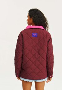 Burgundská prošívaná bunda s růžovým límcem a detaily, se fialovým štítkem značky na zadní straně a uvolněným střihem.