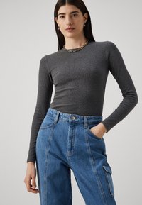 Top grigio a maniche lunghe in cotone con coste abbinato a jeans blu a vita alta, dotati di tasca laterale e dettagli di cucitura evidenti.