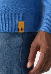 Blauer Strickpullover mit geripptem Saum, versehen mit einem gelben Etikett und einem lila Logo an der Seite. Hand ruht auf Denim-Stoff.