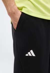 Άτομο που φοράει μαύρο παντελόνι Adidas με το χέρι στην τσέπη και ένα ανοιχτό πράσινο πουκάμισο μερικώς ορατό.