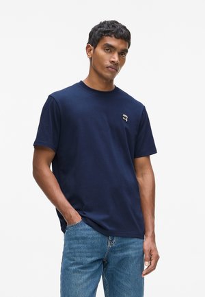 Mężczyzna w granatowej koszulce z małym logo na piersi i niebieskich dżinsach, stojący z jedną ręką w kieszeni na białym tle.