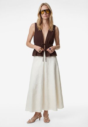 Vrouw met geelgetinte zonnebril, mouwloze bruine top met een knoop aan de voorkant, lange witte textuurrijke rok en beige sandalen met hak.