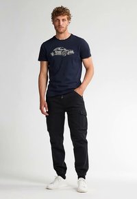 Tmavě modré tričko s grafikou světle zbarveného auta; v kombinaci s černými nákladními kalhotami s kapsami a bílými teniskami. Ležérní střih a design.