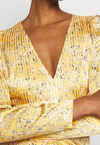 Gul mönstrad blus med djup V-ringning, som har vertikala ränder och en mjuk textur. Långa ärmar med puffdetaljer.