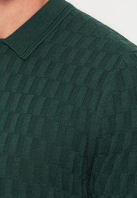 Polo en maille vert foncé avec un motif damier texturé visible sur l'épaule supérieure et le cou d'une personne.